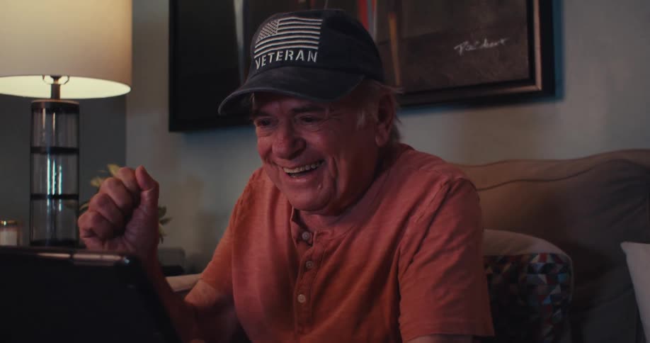 An elderly man wearing a veteran cap smiles with joy while looking at a tablet