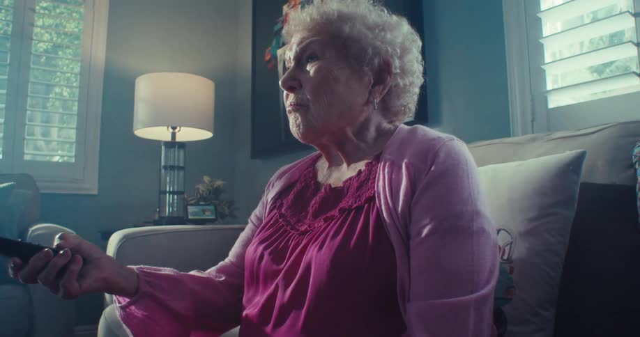 An elderly woman sits on a couch holding a remote looking contemplative in a softly lit living room
