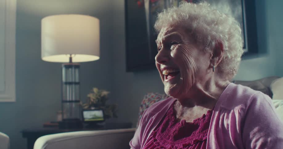 Elderly woman smiles and laughs while sitting on a couch in a living room