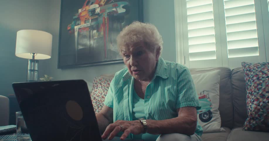 An elderly woman is using a laptop while sitting on a sofa and then she starts smiling