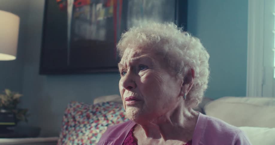 An elderly woman sits alone on a couch tearfully looking away in a quiet emotional moment