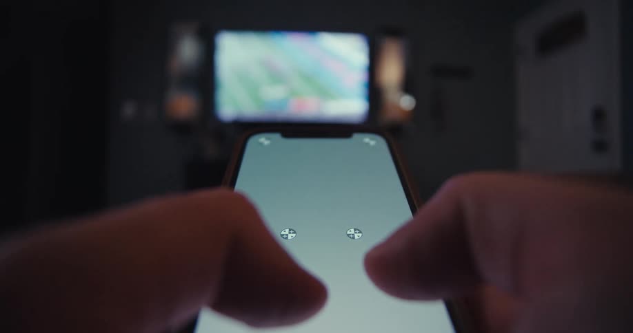 A firstperson perspective shows hands operating a smartphone with a white replaceable screen while a blurred television displaying colorful content is visible in the dark background