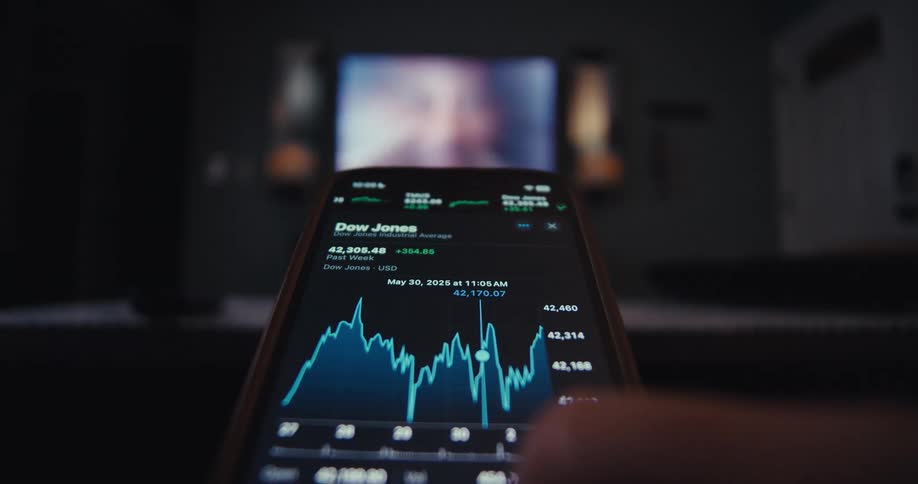 A persons hand holds a mobile device checking stock market prices while sitting in front of a television indoors