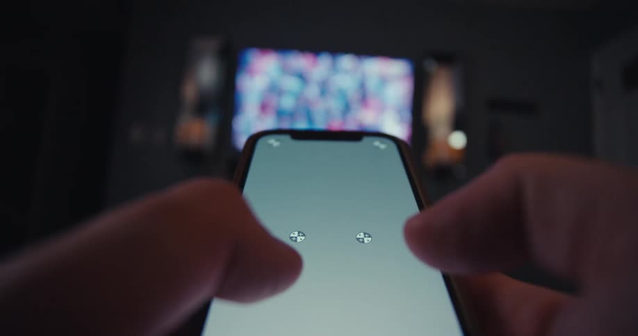 Person uses smartphone as game controller in front of blurred tv in dim room