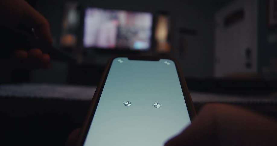 A persons finger touching the screen of a cell phone in an indoor environment with a tv in the background