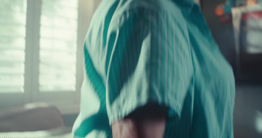 An elderly woman with blue eyes wearing matching turquois blue socks shoes and layered tops waves her arms around and shuffles her feet as she dances in her bright sunlight filled living room