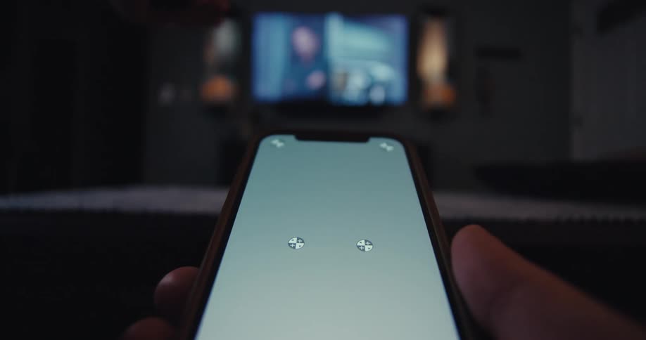 A persons hand holding a cell phone with a gray screen and a tv on in the background