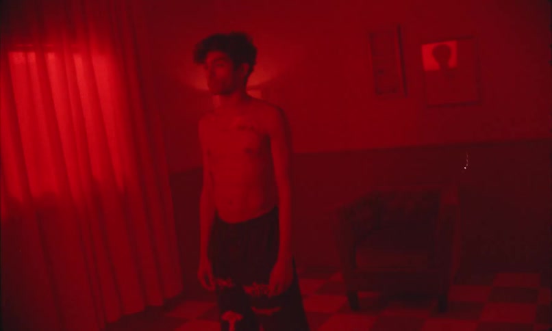 A shirtless young man stands quietly under red light in a dark room