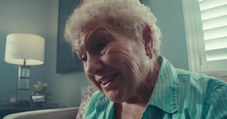 An elderly woman smiles warmly while sitting in a cozy welllit living room