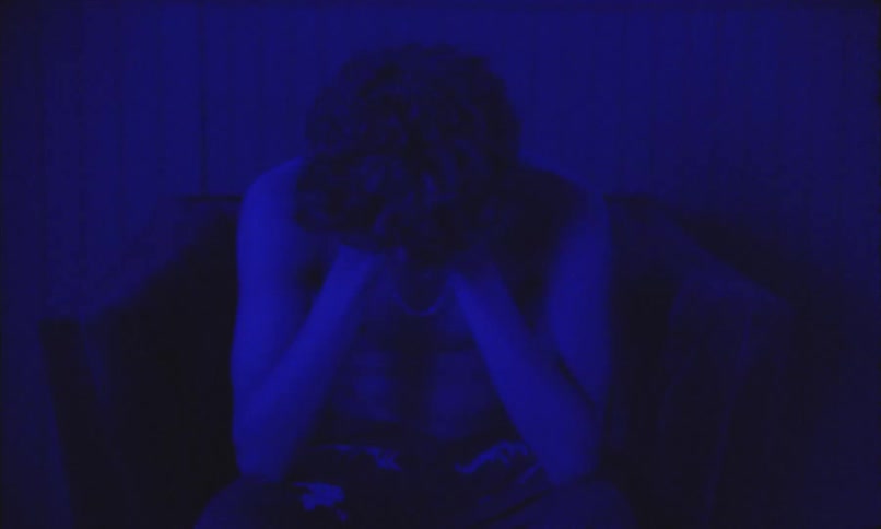 A person sits alone in a dimly lit room with blue lighting head in hands surrounded by checkered flooring a lamp and framed pictures on the wall