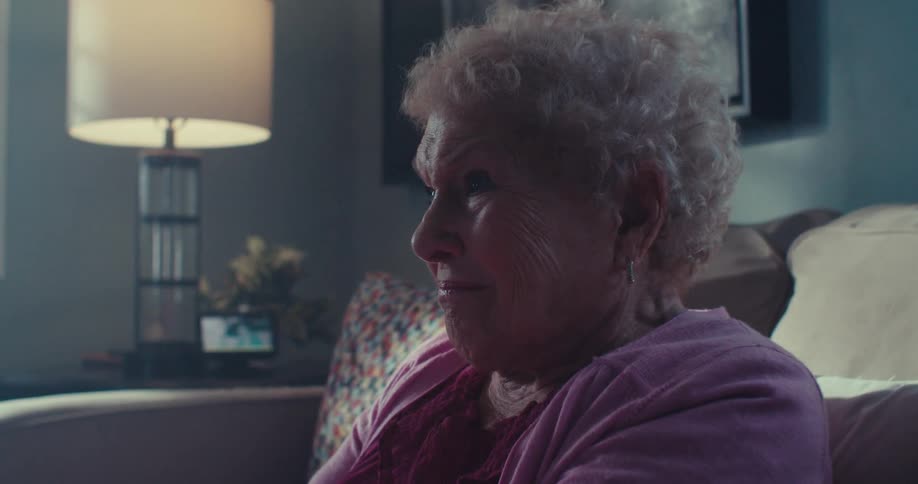 An elderly woman sits on a couch indoors holding a controller and playing a game