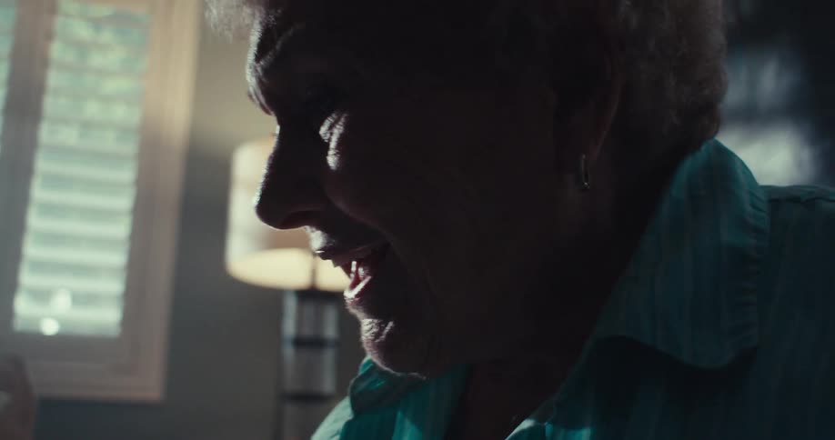 An elderly woman smiles and waves to another person via a tablet computer