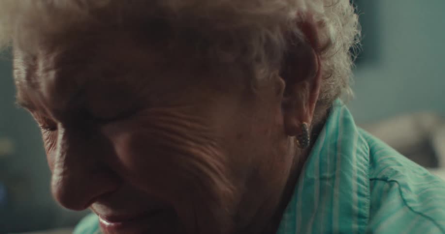 An elderly woman sits on a couch with her hands on her chin smiling