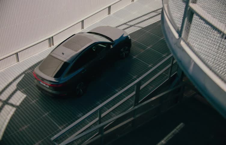 A black car drives along a spiraling metal ramp in a modern urban structure under sunlight