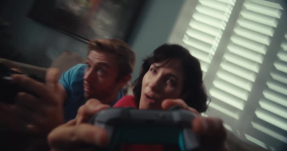 A couple sits on a couch playing video games holding controllers and celebrating with excitement and joy