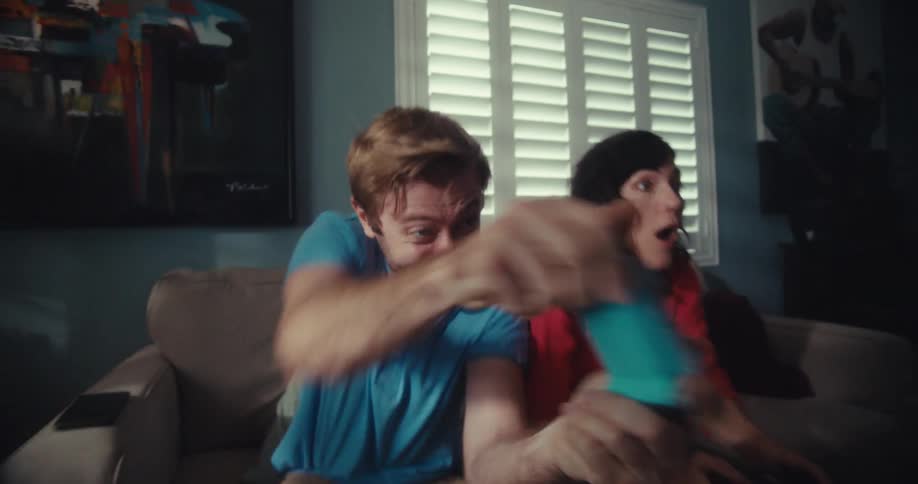 A couple sits on a couch playing video games holding controllers and celebrating with excitement and joy