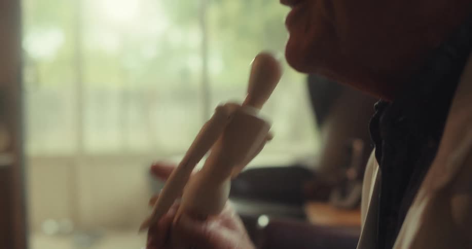 An elderly man wearing a white suit and glasses sits in his office laughing and talking to another person while holding a doll