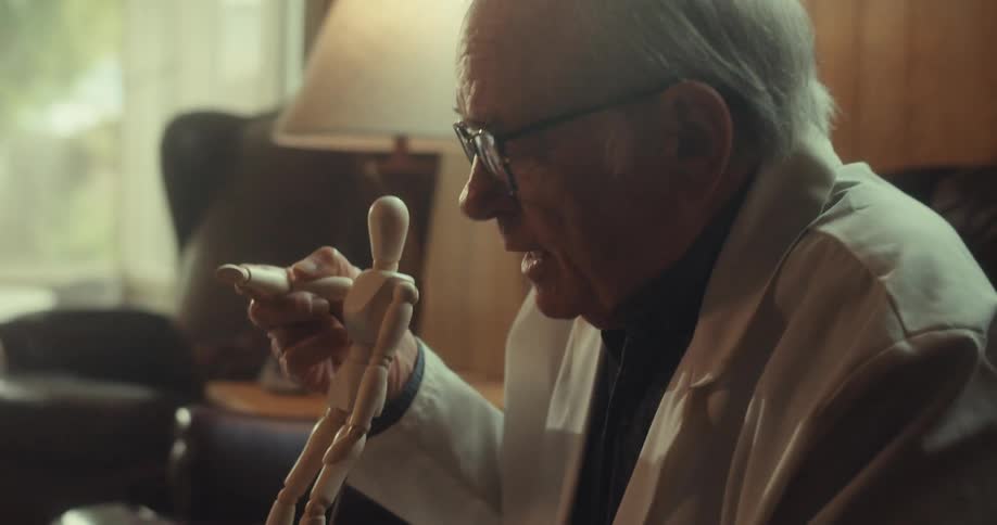 An elderly man in a white coat examines a wooden mannequin in a warmly lit room