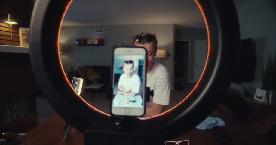 A man films himself with a smartphone mounted on a ring light while wearing sunglasses on his head in a dimly lit room