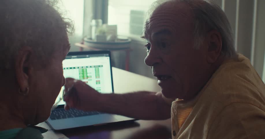 An elderly couple reviews a spreadsheet on a laptop with the man becoming increasingly animated as he explains something before pointing excitedly at the screen shouting with joy and hugging his partner