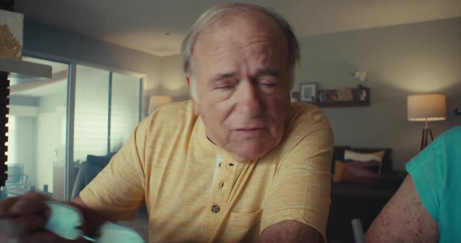 An elderly man sits beside his wife facing a computer putting on his glasses to read whats on the screen while the woman holds a pen and writes something on a piece of paper