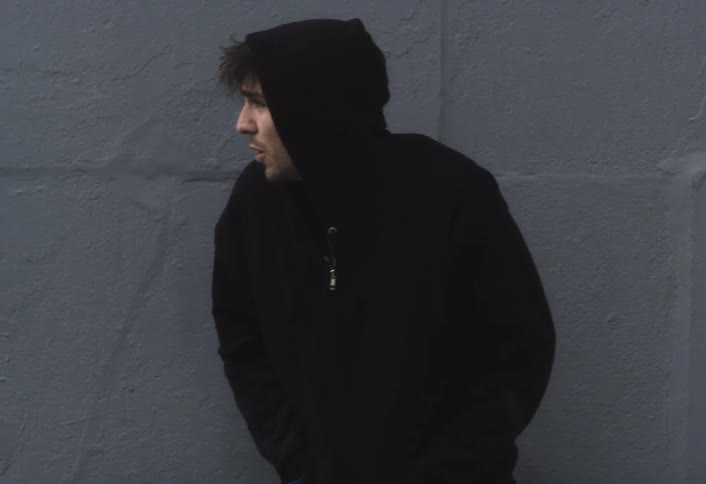 A man runs off the street and stands by a building wall covering his head with his hoodie at midday