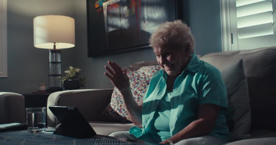 An elderly woman with short blonde hair sits on a couch smiling and waving as she has a video call on a tablet