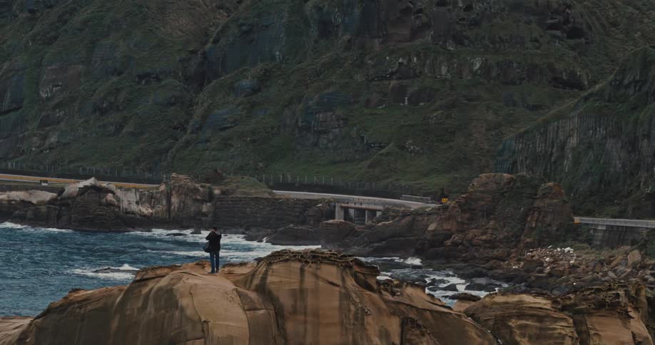 A person photographs a rugged coastal view from a rocky cliff