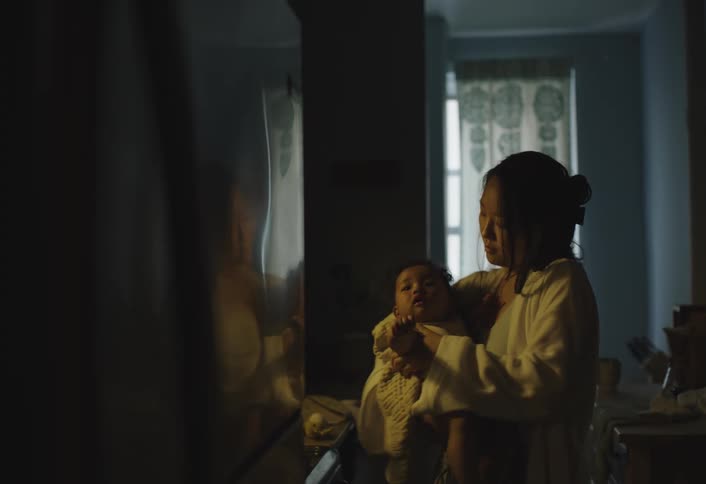 A mother gently holds her baby in a dimly lit room near a window