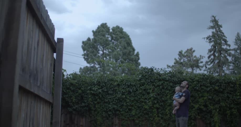 A person stands in a backyard holding a child both gazing upward beneath an overcast sky surrounded by a wooden fence and dense foliage