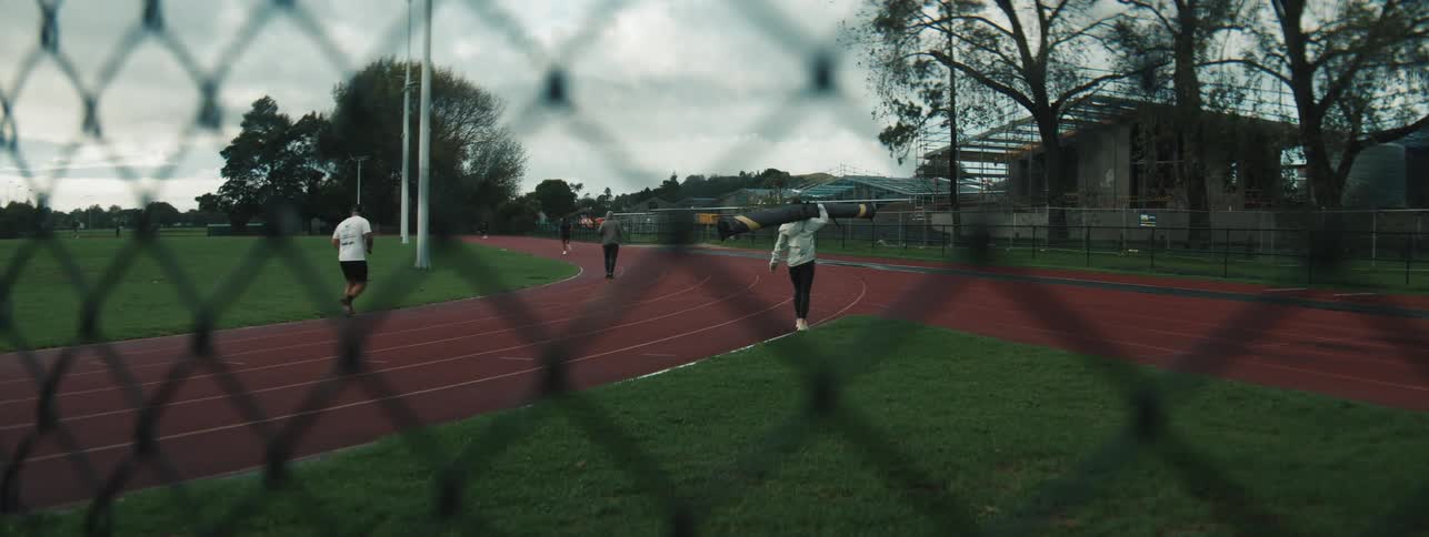 Several people jog and walk along a red running track enclosed by a chainlink fence with trees and buildings in the background