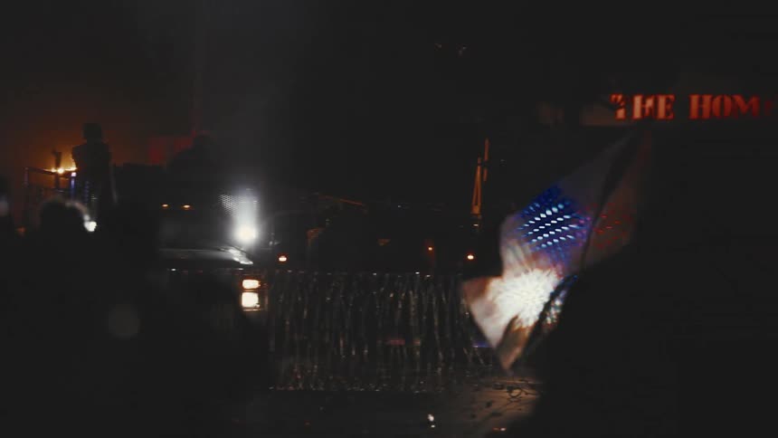 A diverse crowd of protesters gathers on a street at night some holding flags facing several armored vehicles and police cars