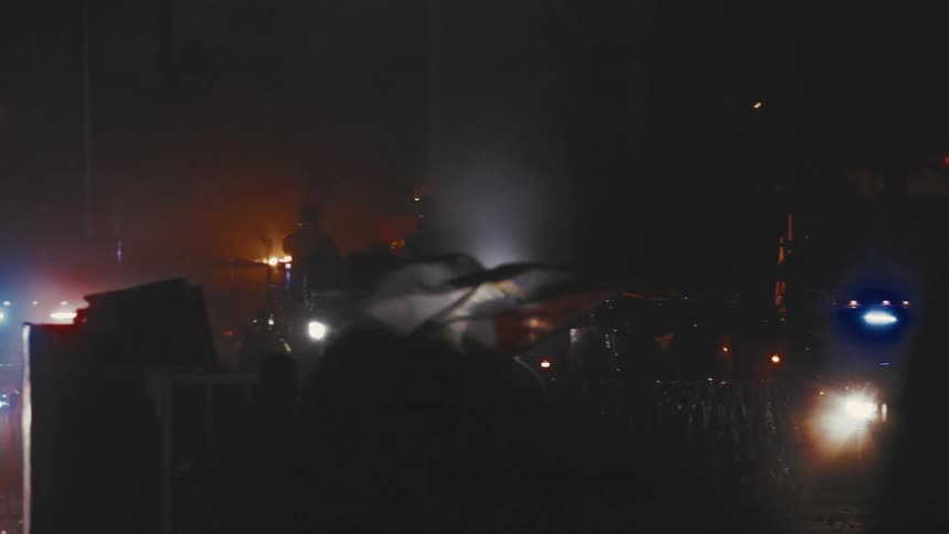 A diverse crowd of protesters gathers on a street at night some holding flags facing several armored vehicles and police cars
