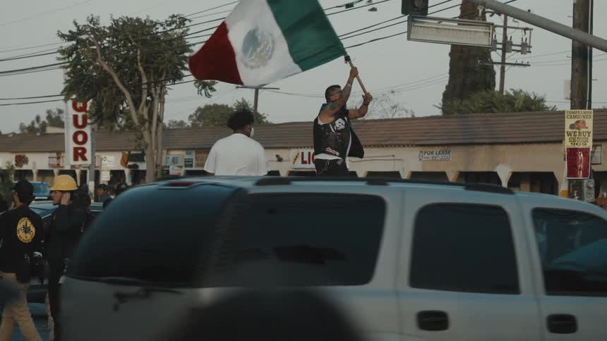 Protesters crowd a street as a man waves a large mexican flag atop a graffiticovered car