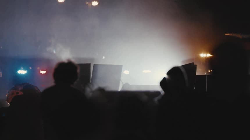 A crowd of protesters gathers on a street at night some holding signs facing several armored vehicles and police cars
