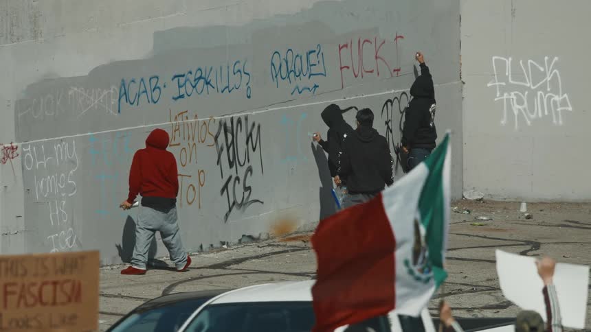 A group of people are spray painting graffiti including slogans insults and political statements on a wall during what appears to be a protest with others holding signs and a flag nearby