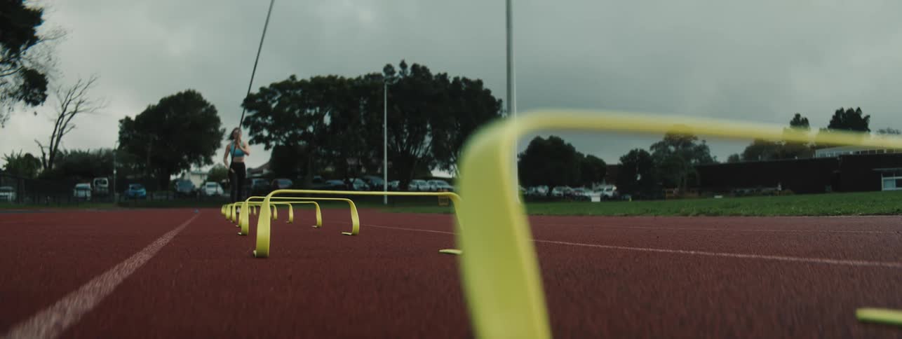 A female athlete runs on a red track while holding a long pole with several yellow hurdles in the foreground and a cloudy sky overhead