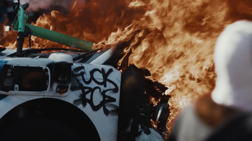 People take video with their smartphones while a car marked with graffiti protesting ice burns