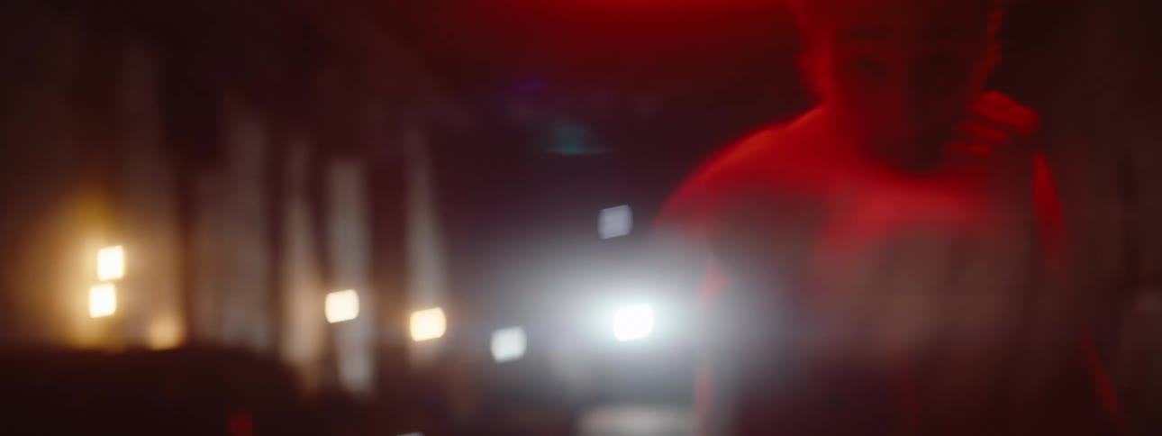 A muscular shirtless man with a beard is boxing on a dark street at night under red lights