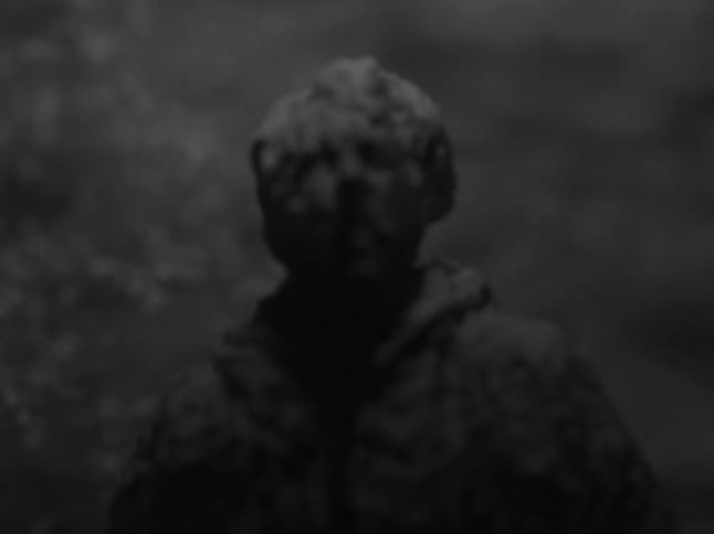 A dark shadowy figure disintegrates into dust against a blurred backdrop of swaying tree branches in black and white