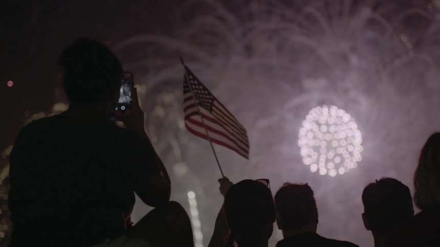 A person waves an american flag in a crowd of people while fireworks explode in the sky