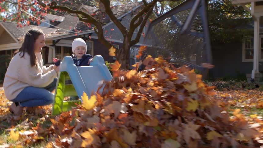 A mother helps her child slide into autumn leaves while the father rakes nearby