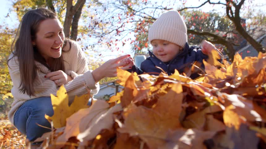 A smiling woman helps a toddler play with autumn leaves in a sunny suburban yard