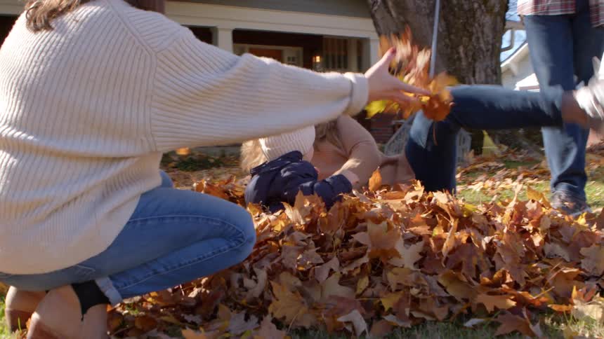 A baby in the middle of many dry leaves with two women nearby throwing more dry leaves all of them having fun