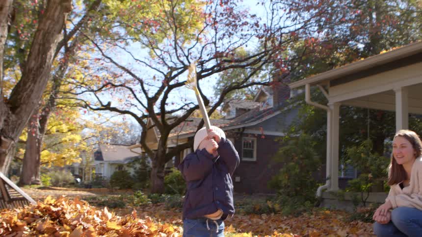 A mother and son play together while the family rakes a large pile of autumn leaves in front of a house on a sunny day