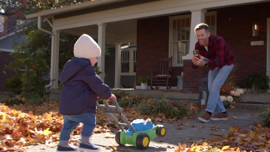 Father films toddler pushing a toy lawnmower on a leafy sidewalk outside a house
