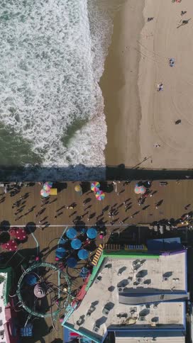 Waves crash along a sandy beach beside a crowded pier lined with people colorful umbrellas and an amusement park