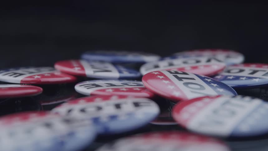 A pile of vote buttons featuring red white and blue designs with stars are scattered on a dark surface
