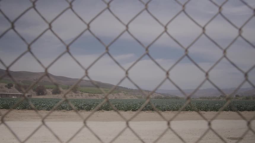 A clear chainlink fence occupies the foreground looking out onto a vibrant green field a dirt path and distant mountains under a cloudy sky