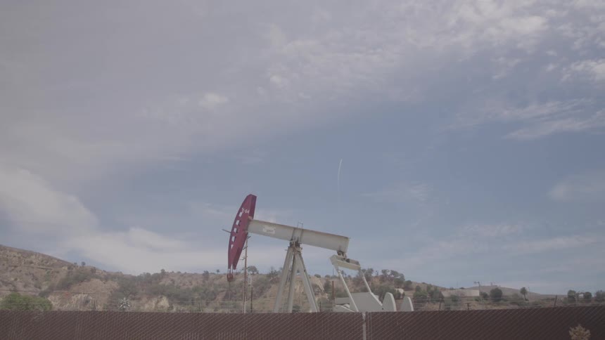 A lone red and white oil pumpjack operates in a dusty landscape under a cloudy sky with distant hills in the background